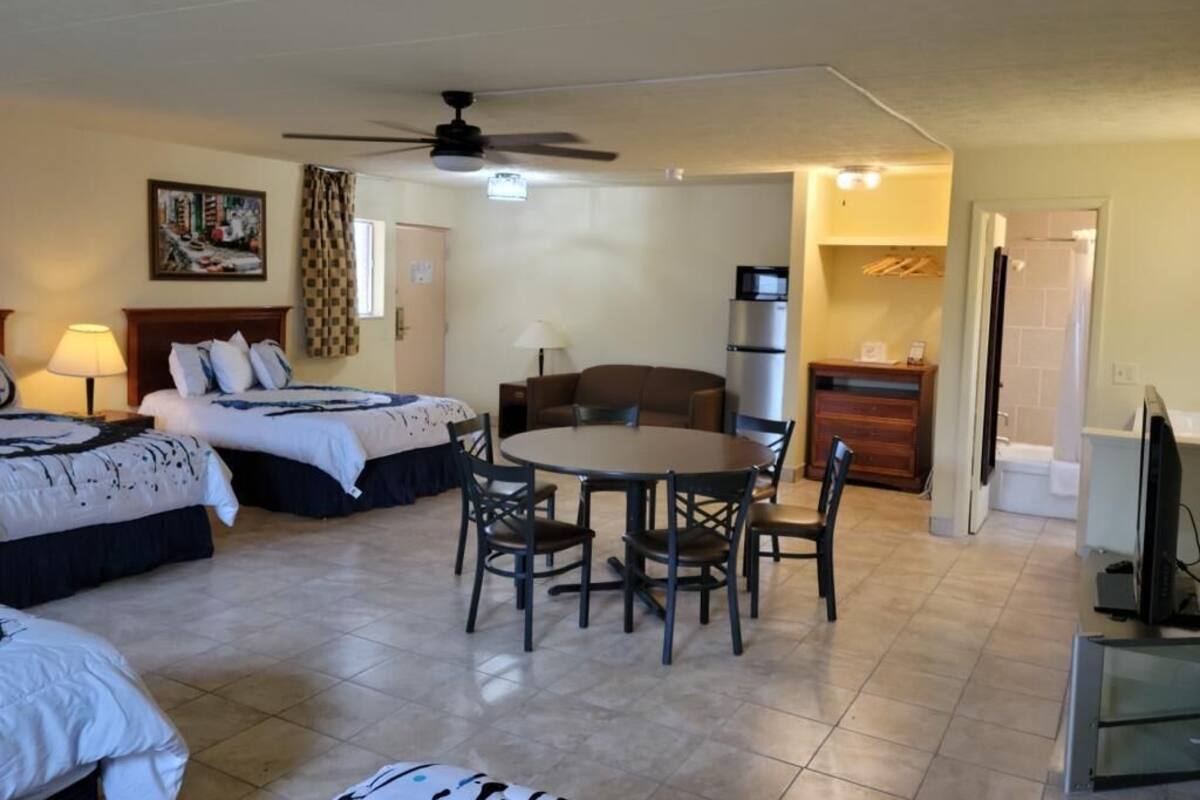 A hotel room with two beds, a dining table, chairs, a ceiling fan, a refrigerator, and a television.