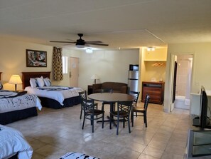 A hotel room with two beds, a dining table, chairs, a ceiling fan, a refrigerator, and a television.