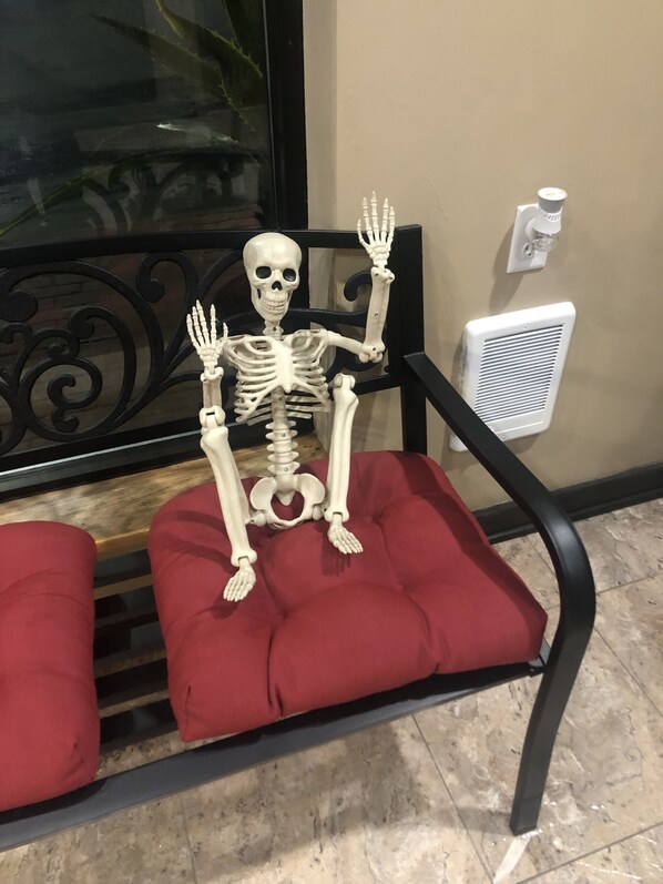 A skeleton seated on a red cushioned chair with a black metal frame, positioned in a room with a tiled floor and a window with a decorative iron grill.