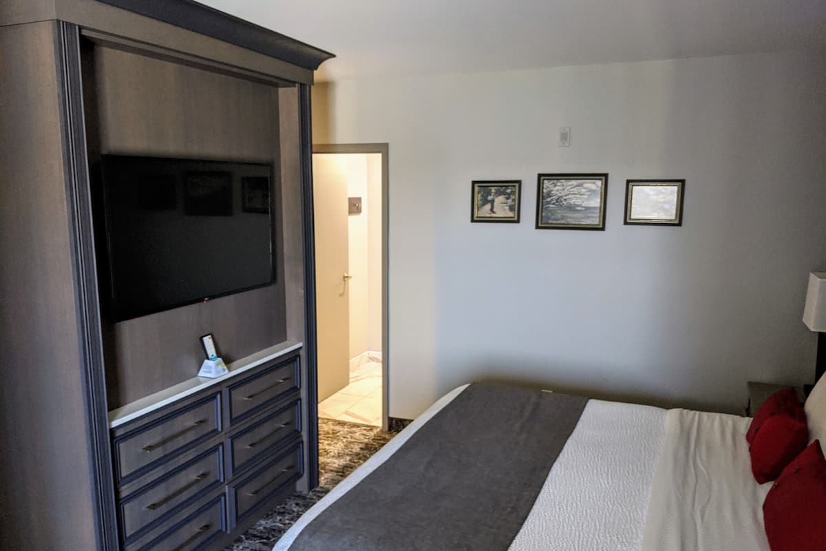 A bedroom with a large flat-screen TV mounted on a dark wooden cabinet, a bed with white bedding and red pillows, and three framed pictures on the wall.