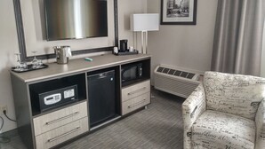 Hotelværelse med fladskærms-TV, skrivebord med kaffemaskine, mikroovn, minikøleskab, varmeblæser og en mønstret lænestol.