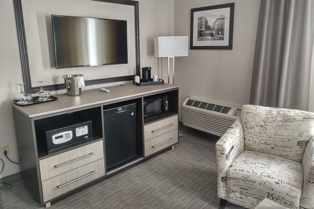 A hotel room with a flat-screen TV, a desk with a coffee maker, a microwave, a mini-fridge, a heater, and a patterned armchair.