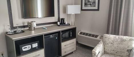 Habitación de hotel con televisión de pantalla plana, escritorio con cafetera, microondas, refrigerador pequeño, calefacción y un sillón estampado.