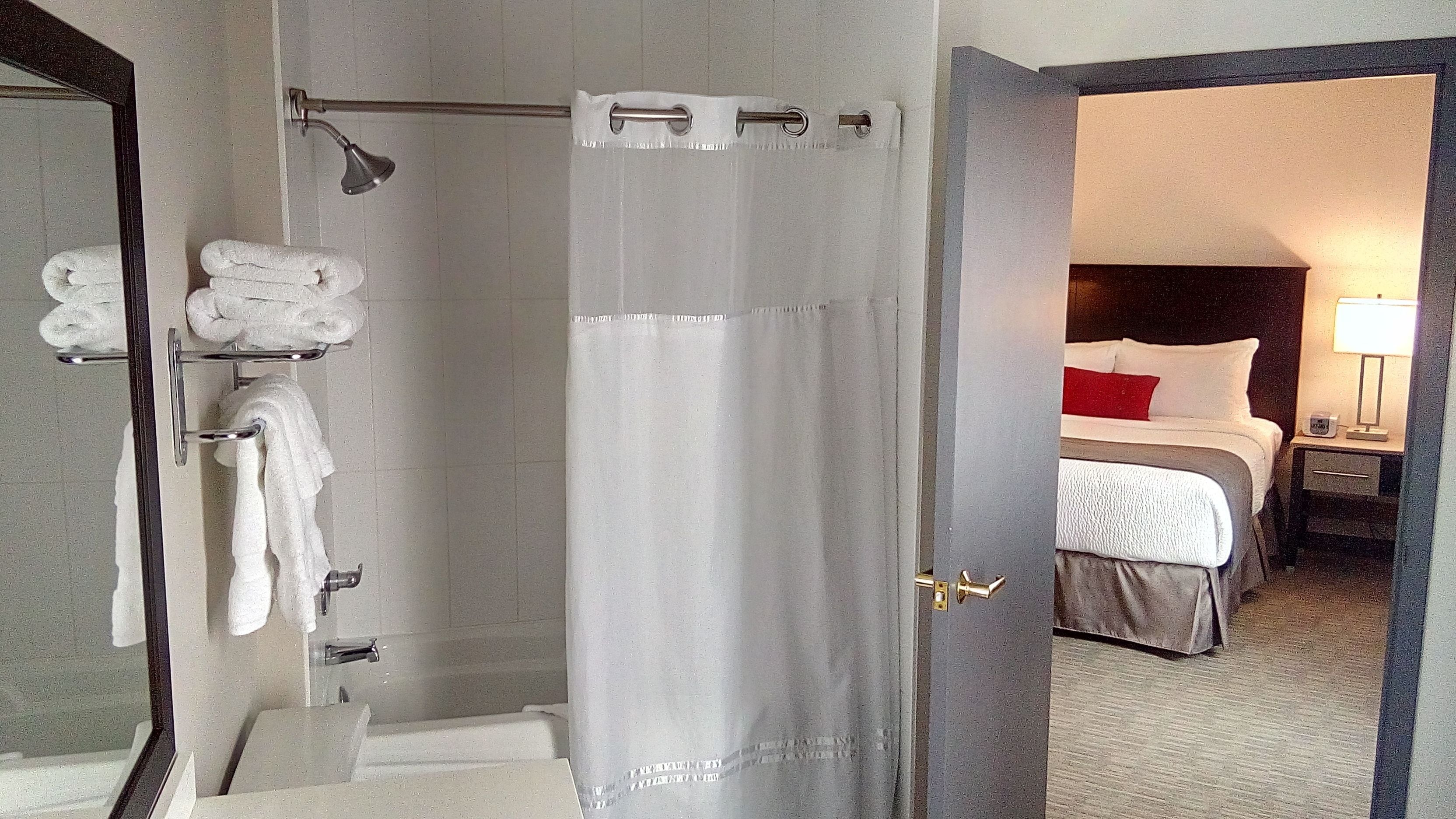 A hotel bathroom with a shower, a towel rack with folded towels, and a visible door leading to a bedroom with a bed, nightstand, and lamp.