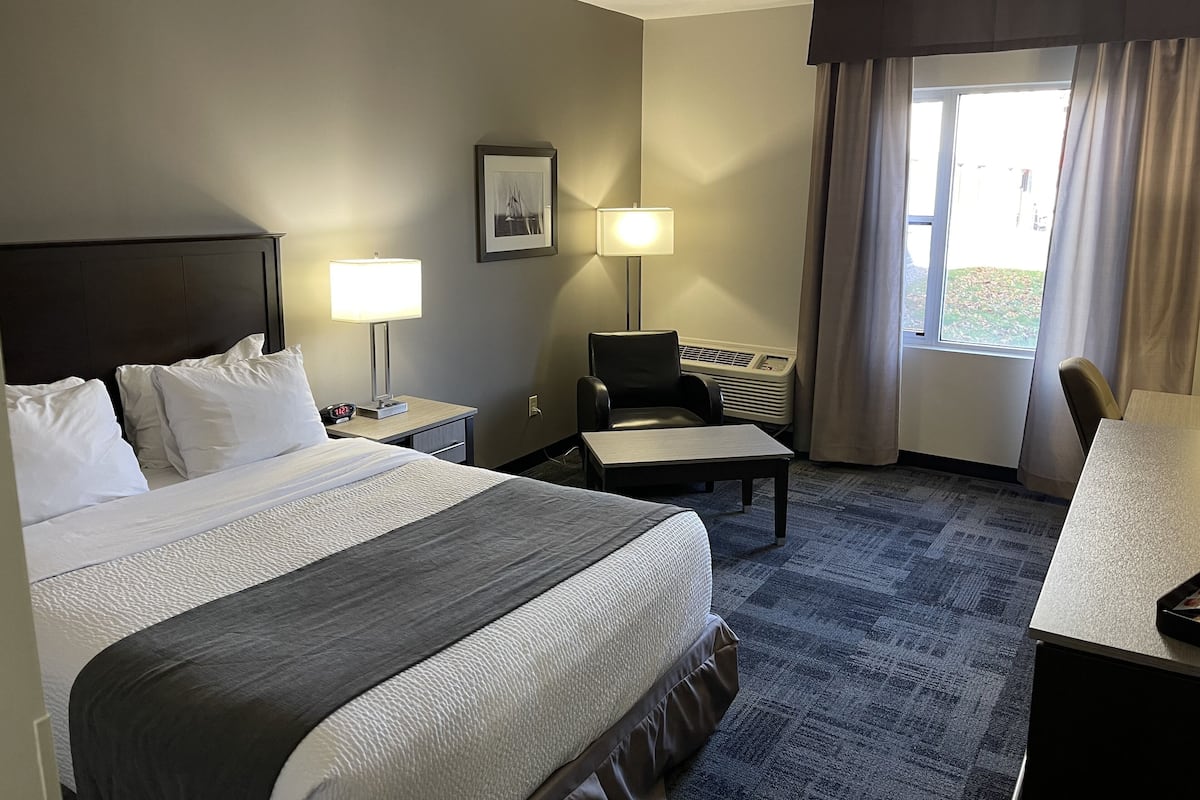 A hotel room with a large bed, a desk, a chair, a lamp, an air conditioner, and a window with curtains.