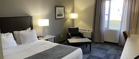 Habitación de hotel con una cama grande, un escritorio, una silla, una lámpara, aire acondicionado y una ventana con cortinas.