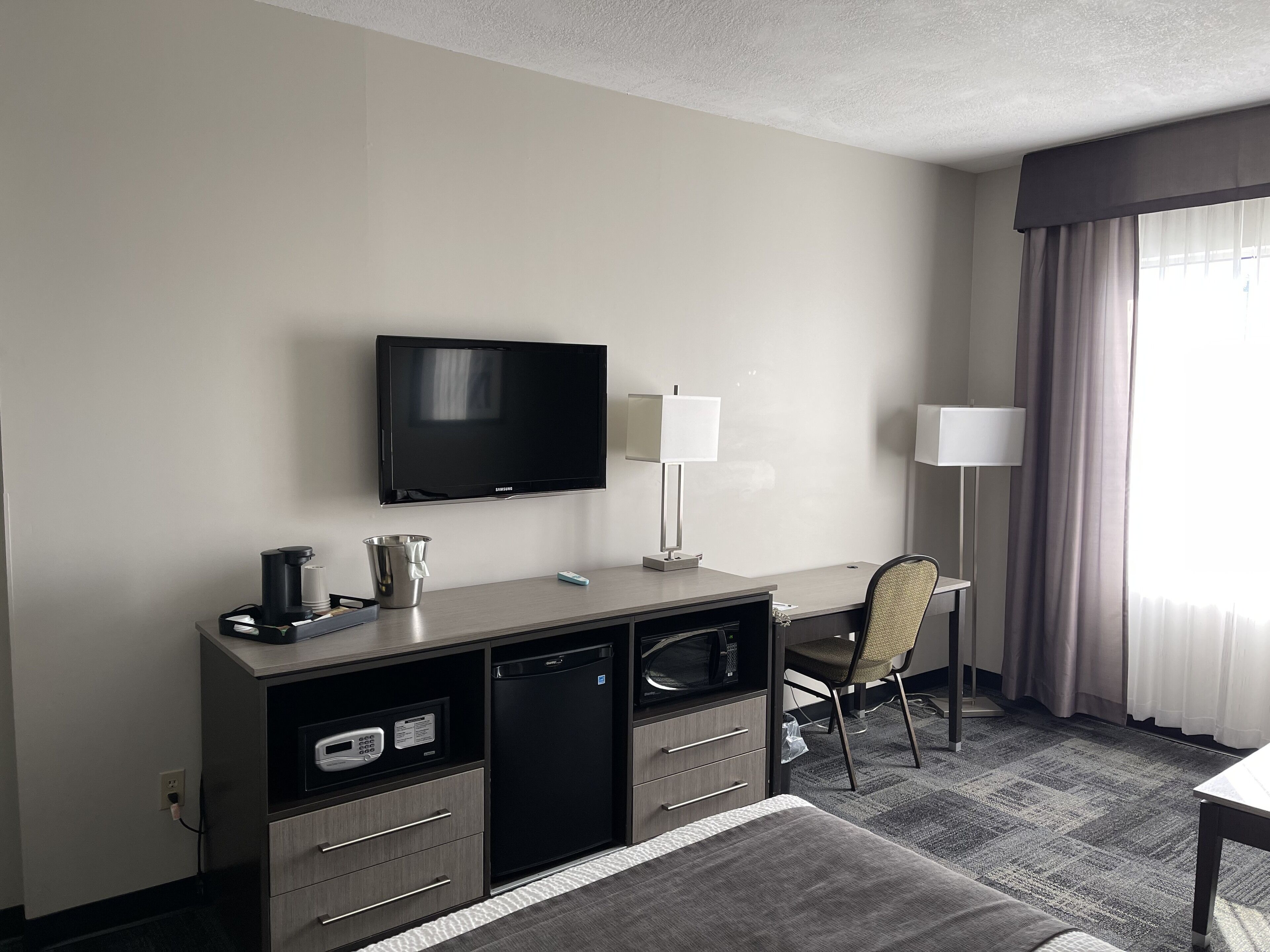 A hotel room with a flat-screen TV, a desk with a lamp, a microwave, a mini-fridge, and a chair.