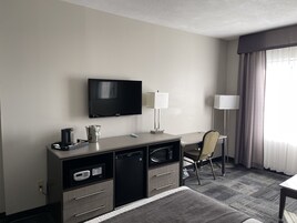 A hotel room with a flat-screen TV, a desk with a lamp, a microwave, a mini-fridge, and a chair.