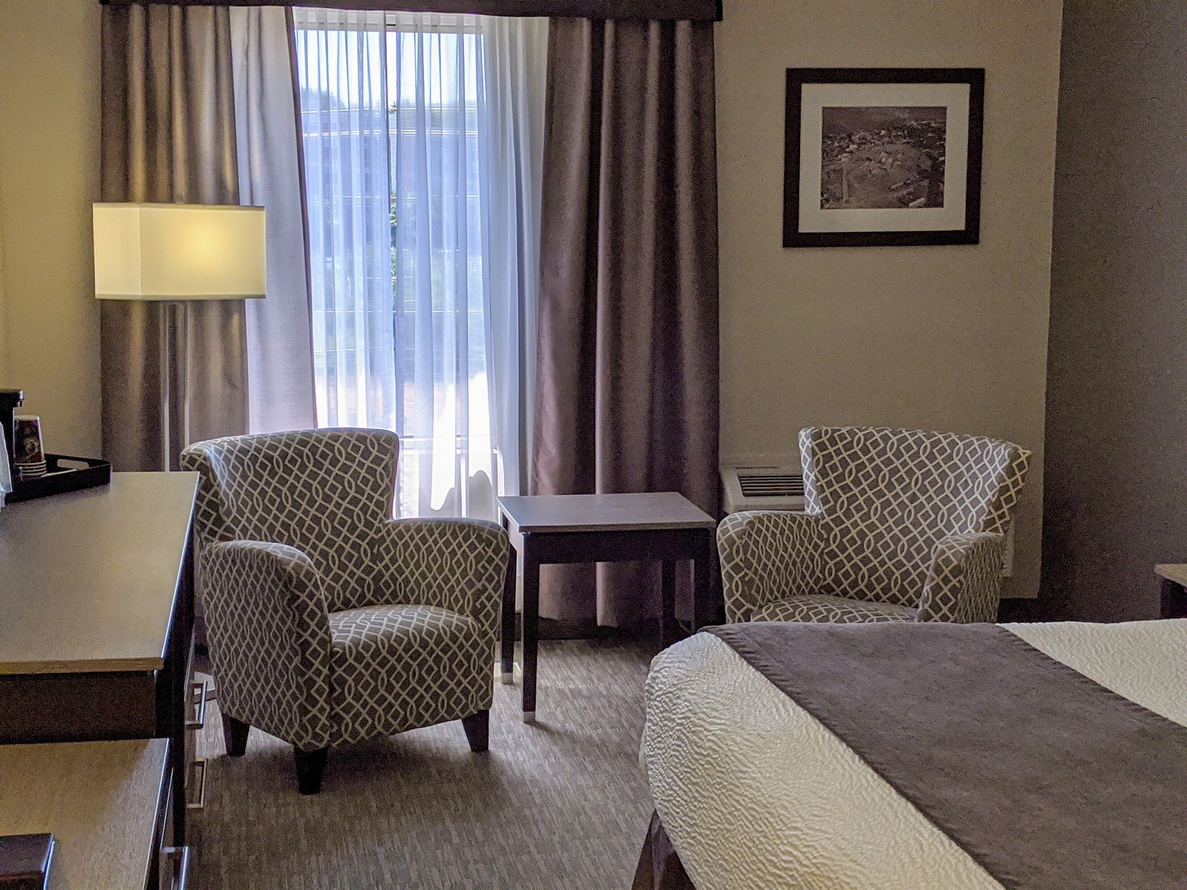 A hotel room with two patterned armchairs, a small table, a bed with a textured blanket, a lamp, and a framed picture on the wall.