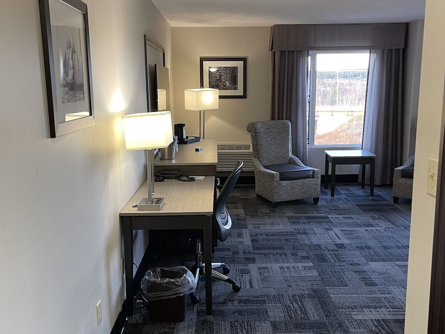 Habitación de hotel con escritorio, silla, lámpara y un amplio ventanal con cortinas.