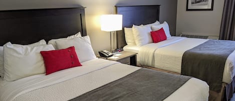 A hotel room with two beds, each with white bedding and red pillows, a bedside table with a lamp, and a framed picture on the wall.