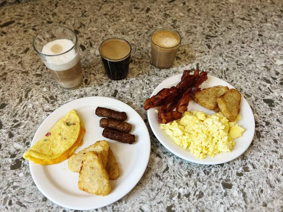 En frukosttallrik med ägg, bacon, korv och rostat bröd, och två glas kaffe på en bänkskiva av granit.