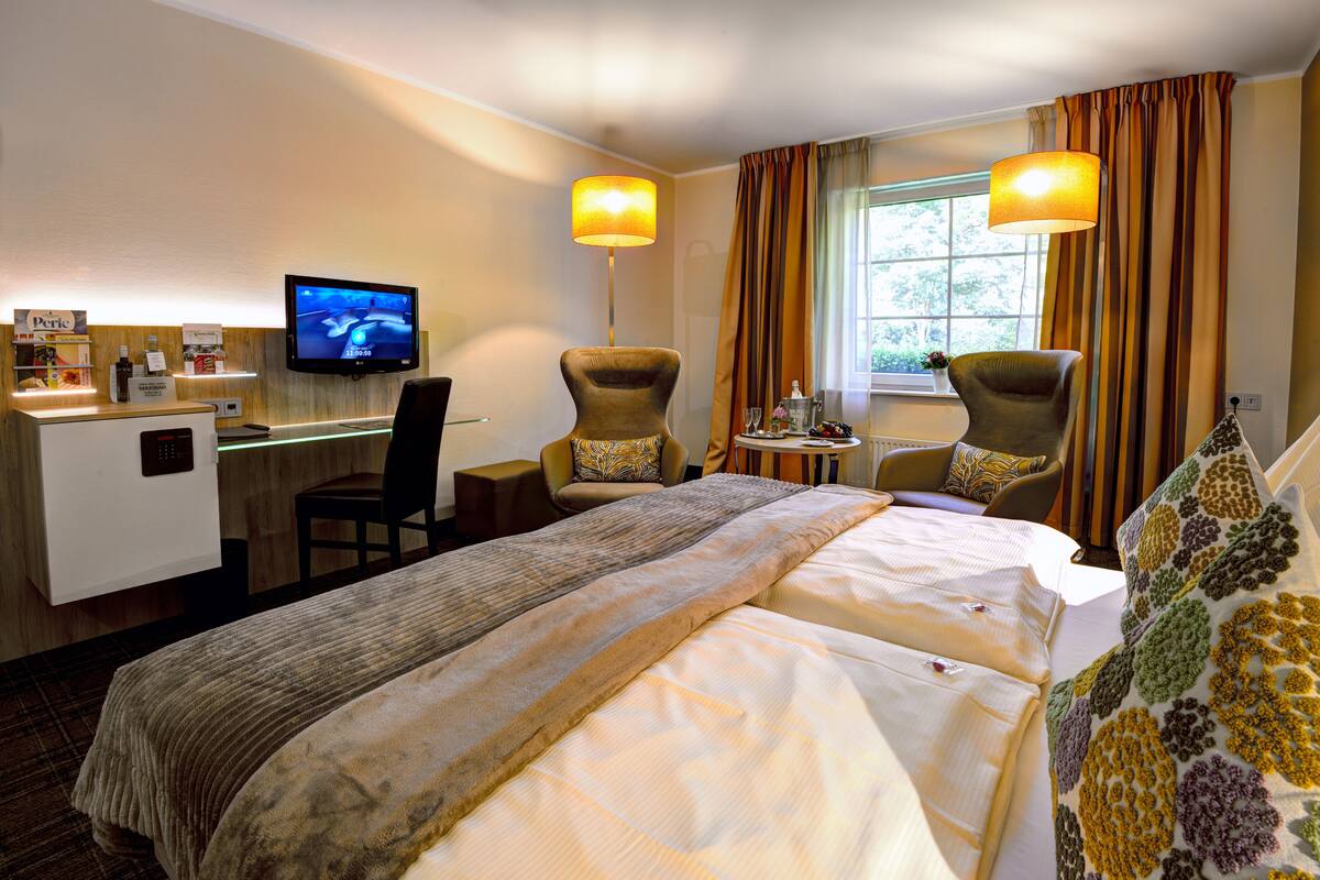 Une chambre d’hôtel comprenant un lit, deux fauteuils, un bureau avec une télévision et une fenêtre avec des rideaux.