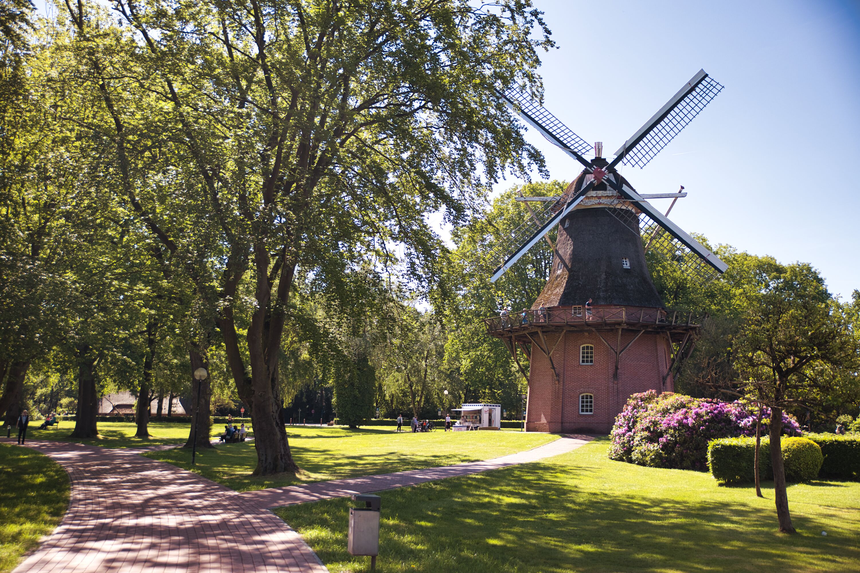 A brick windmill surrounded by lush green trees and a paved walkway.