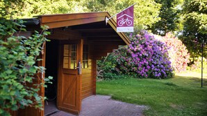Une cabane en bois ornée d’un symbole de vélo, entourée d’une végétation luxuriante et d’arbustes en fleurs.