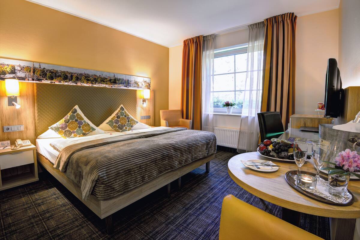 Une chambre d’hôtel avec un lit, une table de chevet, une lampe, une fenêtre avec des rideaux et une petite table sur laquelle se trouve un plateau avec des fruits et des verres.