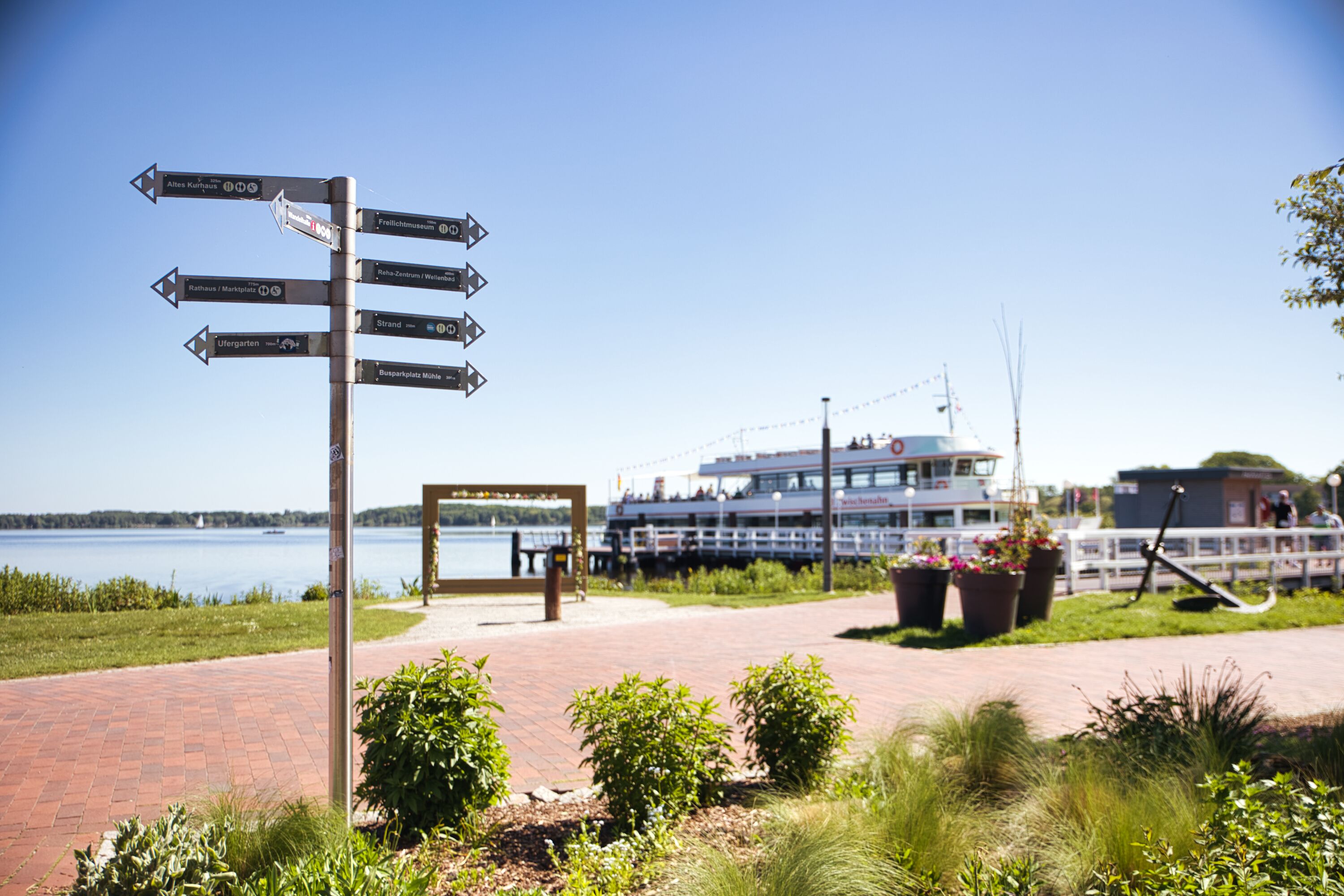A signpost with directions to various locations, a boat docked by the water, and a brick pathway.