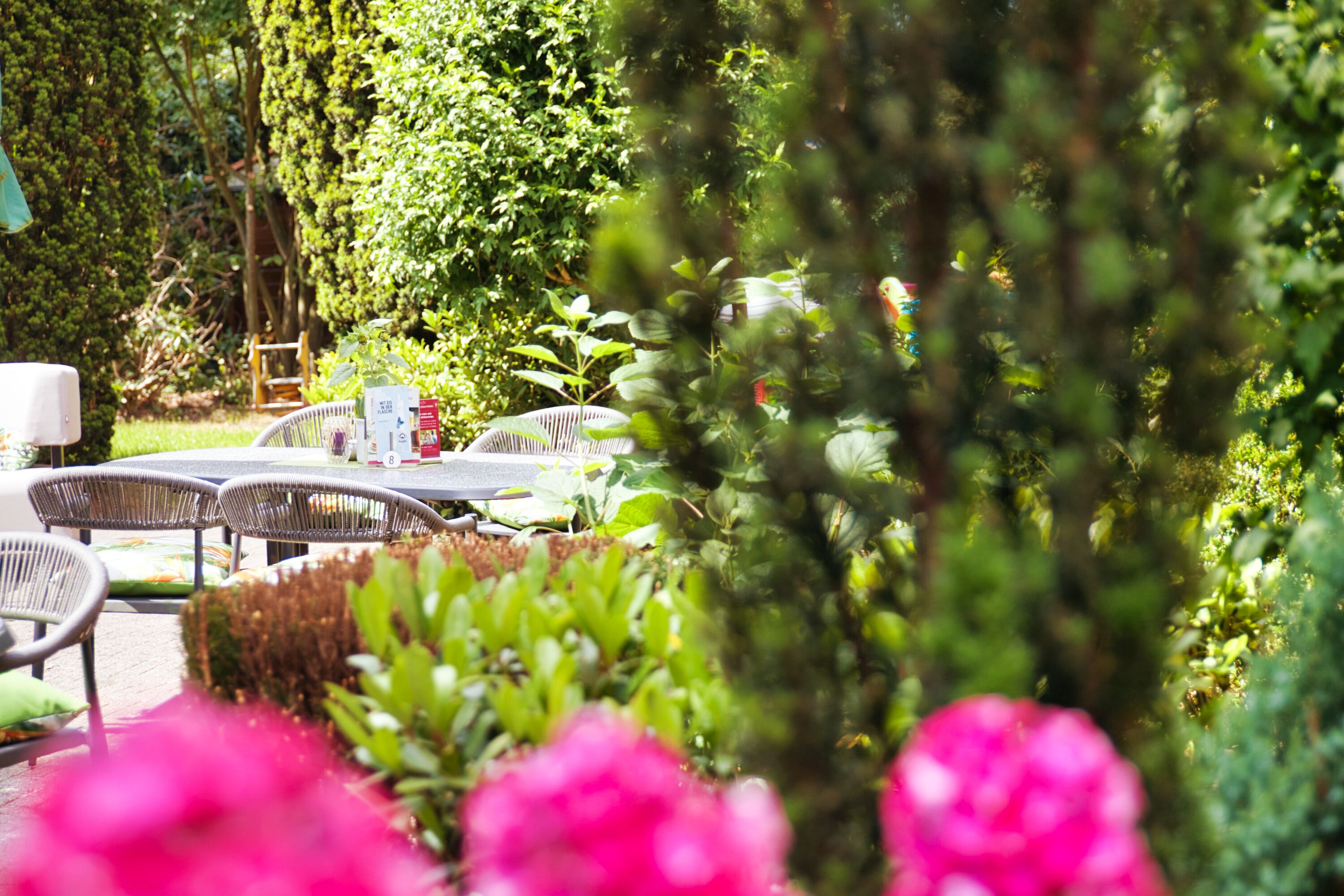 An outdoor seating area with wicker tables and chairs, surrounded by lush greenery and flowering plants.