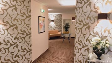 Un corridoio con carta da parati decorata, un quadro appeso, un divano, un tavolo con fiori e un cartello di uscita.