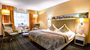 Une chambre d’hôtel avec un grand lit, deux fauteuils, une petite table avec un vase et un tableau représentant un paysage urbain accroché au mur.