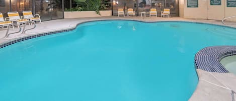 Una piscina interior con agua cristalina, rodeada de sillas de descanso y un muro de vidrio.