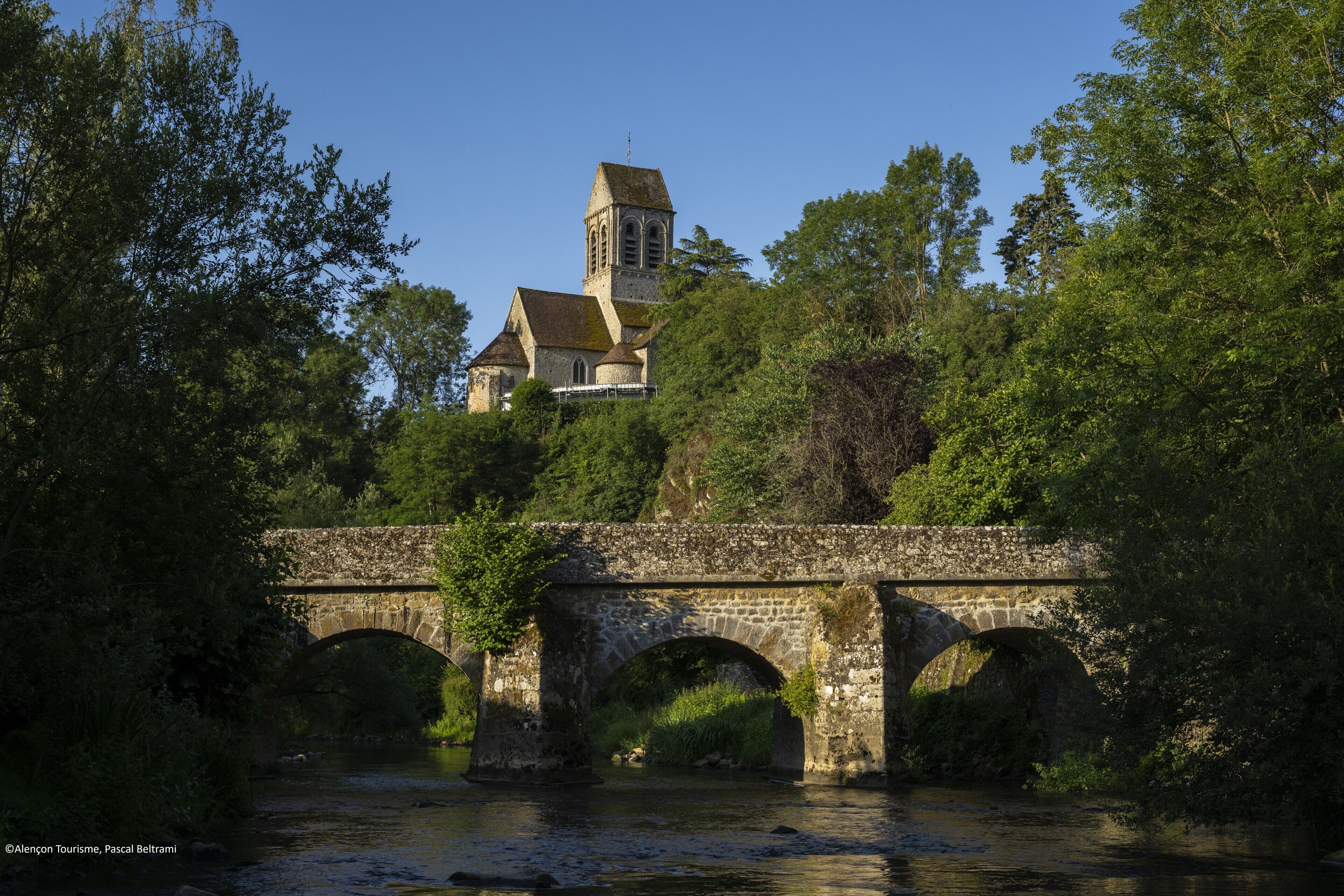 A stone bridge with three arches over a river, leading to a historic building with a tower.