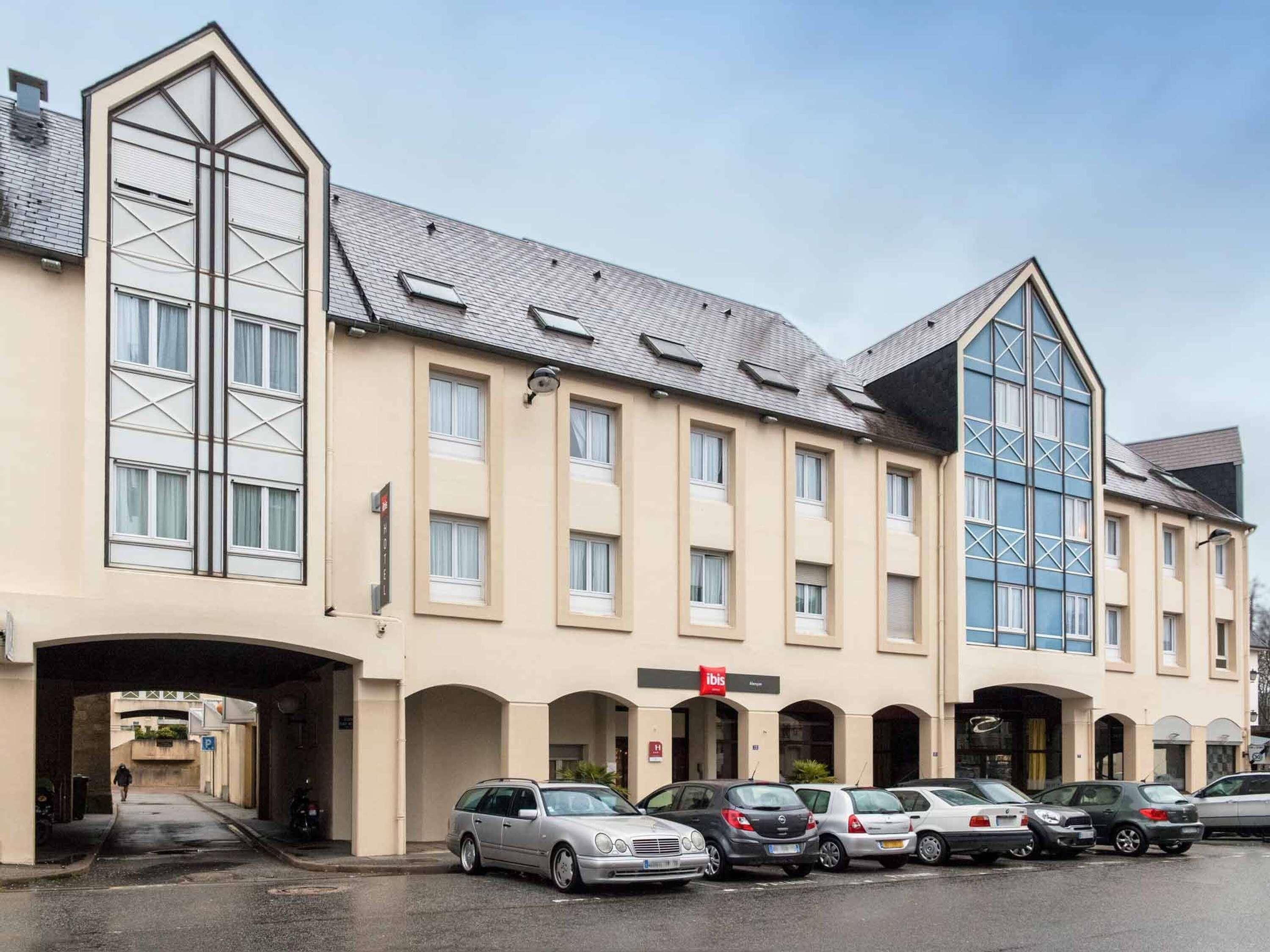 Un hotel con un diseño arquitectónico moderno, que cuenta con una amplia entrada central, varios espacios de estacionamiento y un llamativo frente de vidrio.
