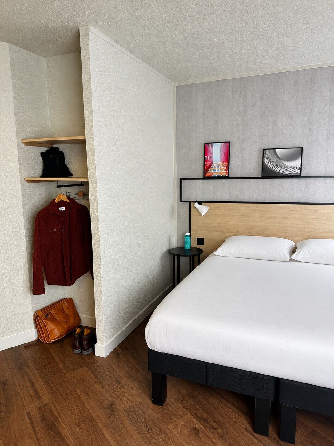 A hotel room with a bed, a wardrobe, a shelf with clothing, and two wall-mounted picture frames.
