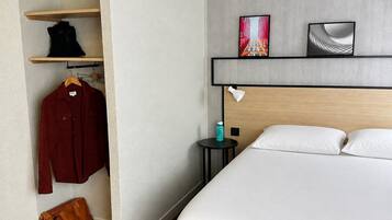 A hotel room with a bed, a wardrobe, a shelf with clothing, and two wall-mounted picture frames.