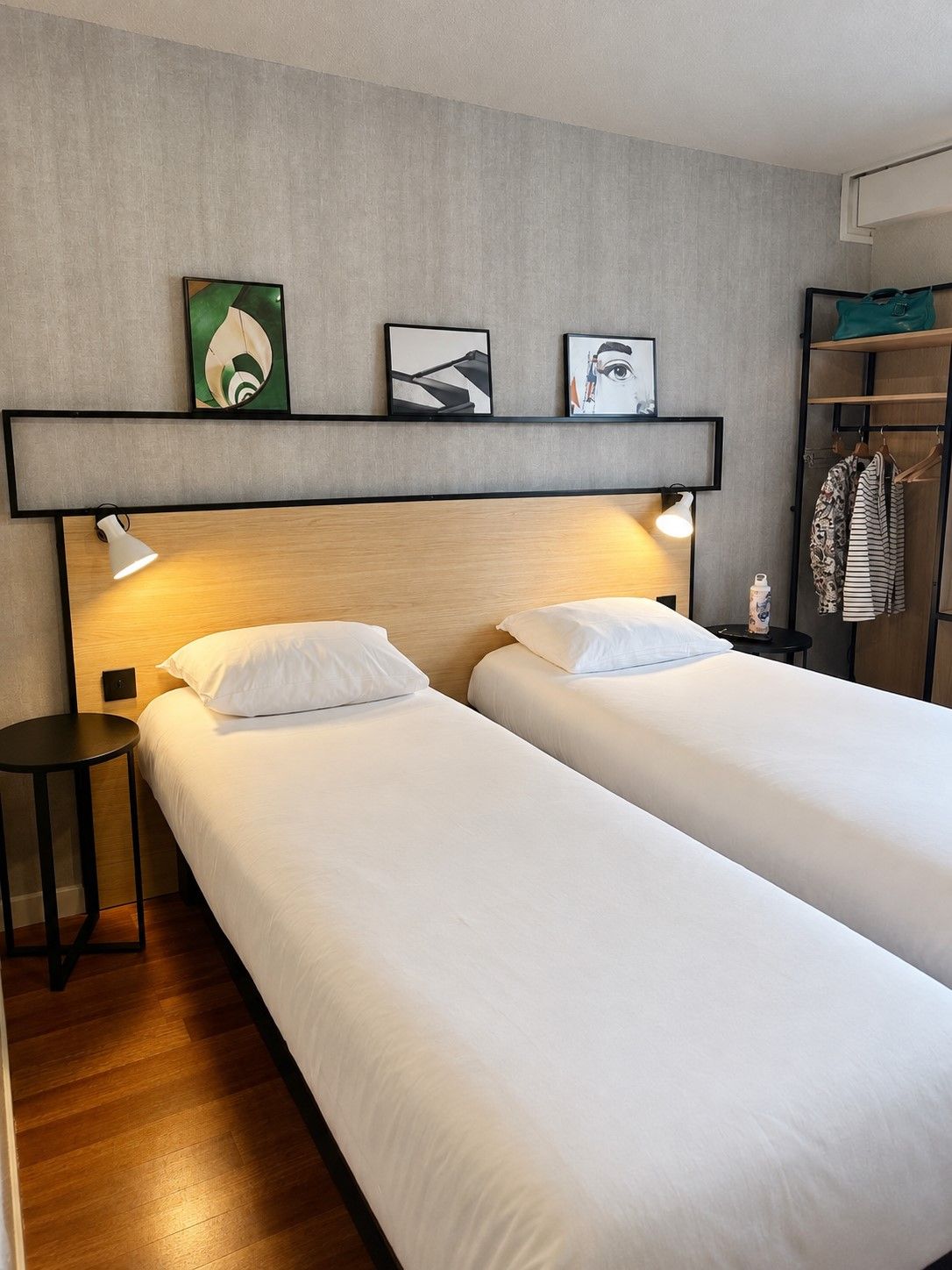Una habitación de hotel con dos camas, una mesita de noche, un estante con obras de arte y un armario.
