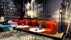 A modern lounge area with a red sofa, blue chairs, and a guitar. - ibis Alencon (Alencon)