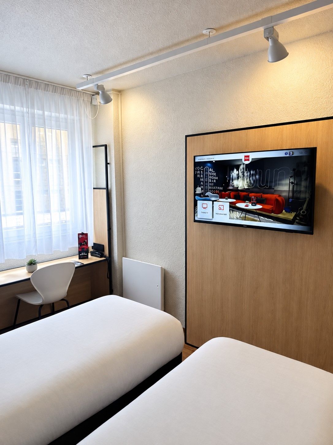 Ett hotellrum med en säng, ett skrivbord, en stol, en tv som är monterad på väggen och ett fönster med gardiner.