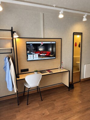 A modern hotel room with a wooden desk, a white chair, a flat-screen TV mounted on the wall, and a mirror reflecting the room.