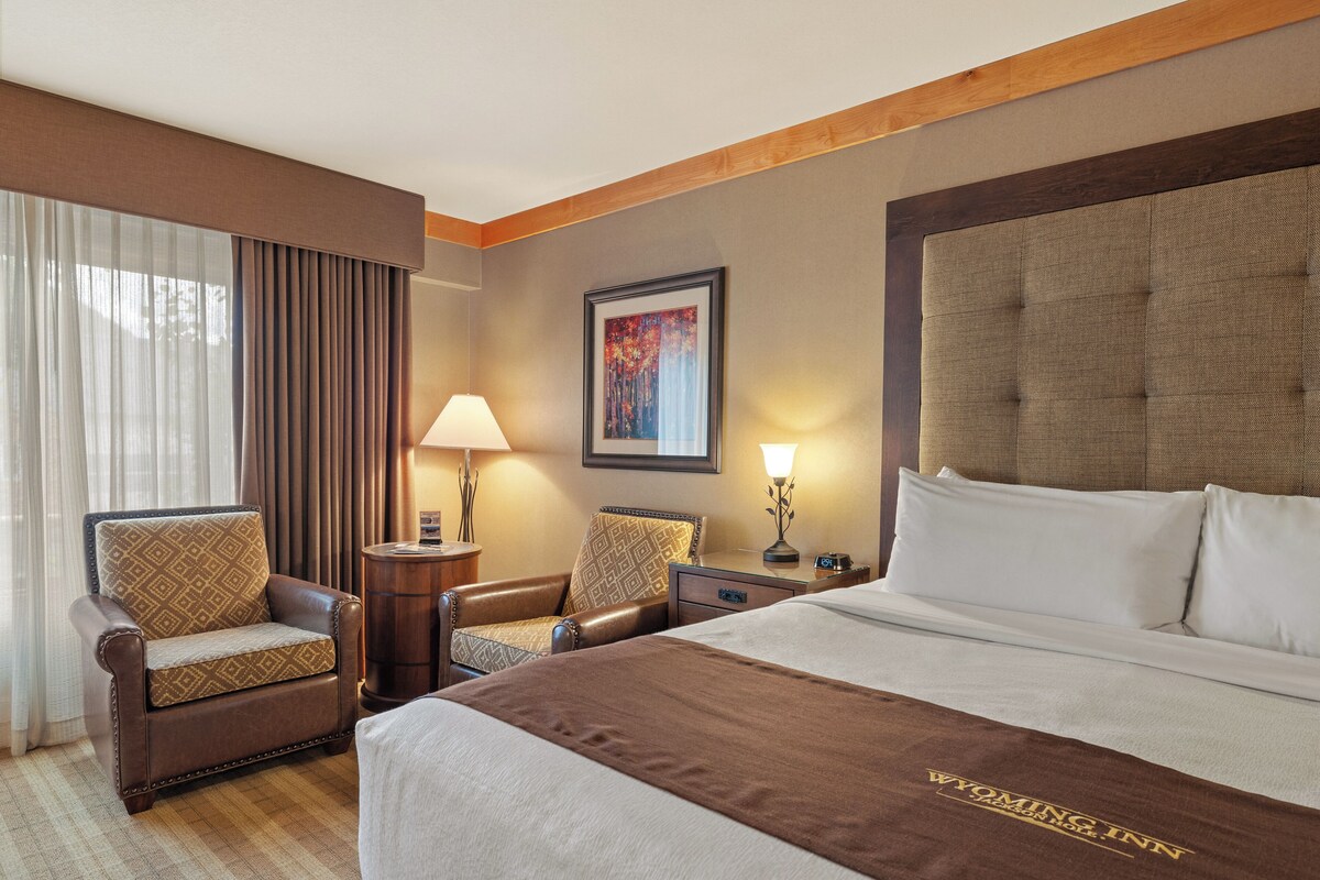 Un cuarto de hotel con una cama grande, dos sillones, una mesita de noche con lámpara y un cuadro en la pared.