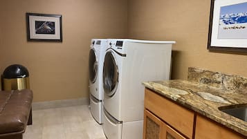 A laundry room with two white washing machines, a brown bench, a sink with a granite countertop, and framed pictures on the walls.