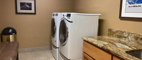 A laundry room with two white washing machines, a brown bench, a sink with a granite countertop, and framed pictures on the walls.