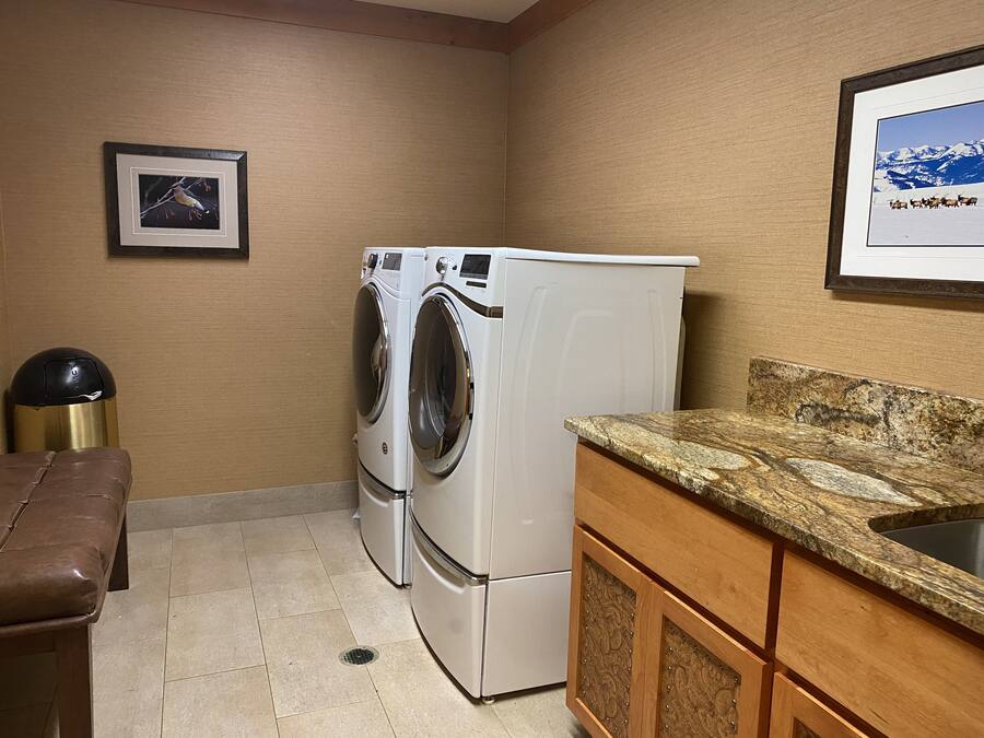 A laundry room with two white washing machines, a brown bench, a sink with a granite countertop, and framed pictures on the walls.
