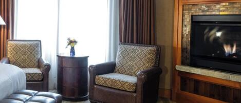 Une chambre d’hôtel avec un canapé en cuir, deux fauteuils à motifs, une petite table d’appoint avec un vase, une cheminée et un grand écran de télévision.