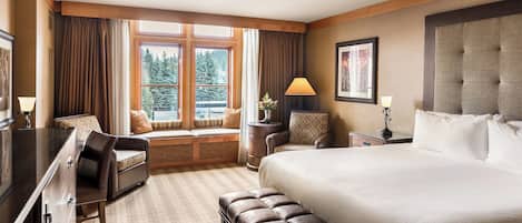 A hotel room with a large bed, a desk, a chair, a window with a view of trees, and a lamp.