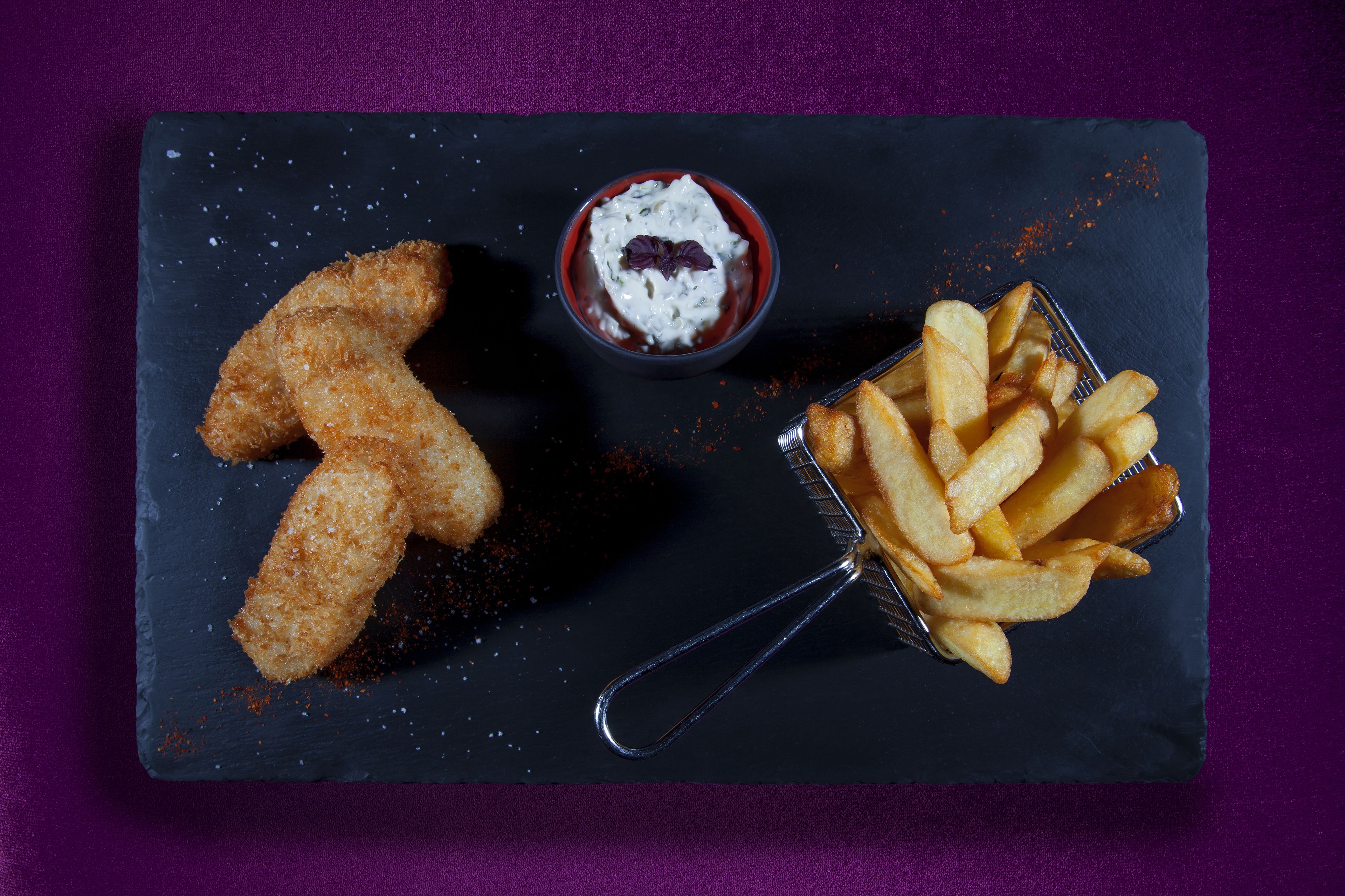 A black slate serving board with breaded chicken tenders, a small bowl of sauce, and a basket of French fries.