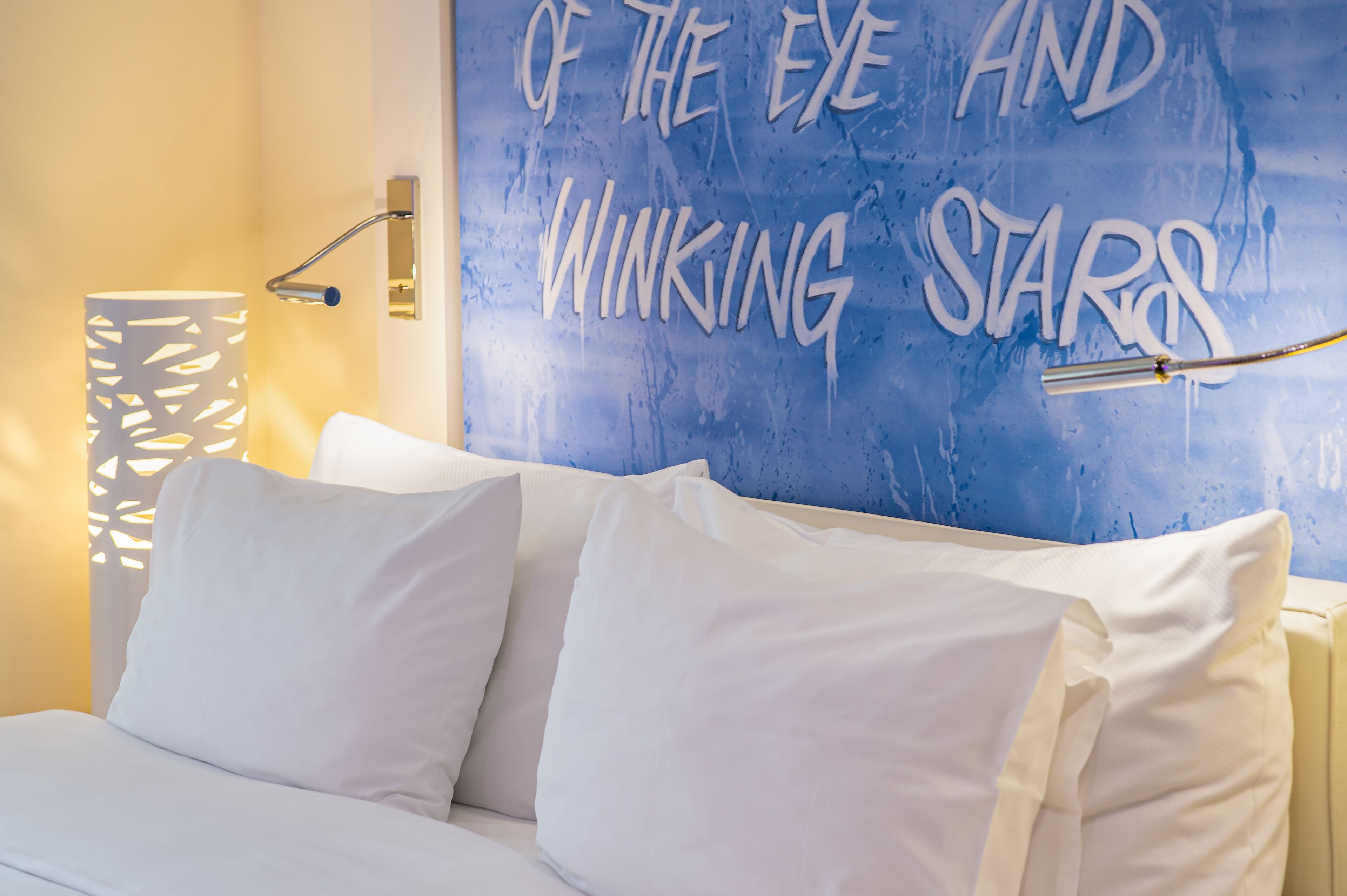 A bed with white bedding and a wall with a blue textured design and the phrase "OF THE EYE AND WINKING STARS".