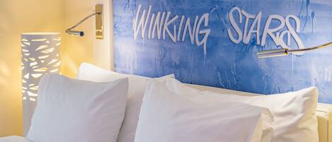 A bed with white bedding and a wall with a blue textured design and the phrase "OF THE EYE AND WINKING STARS".