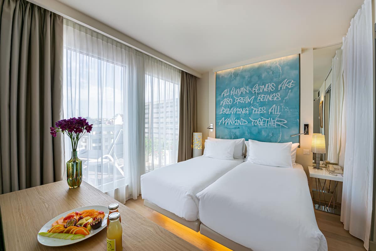 A hotel room with two beds, a dining table with breakfast, a vase of flowers, and a motivational quote on the wall.
