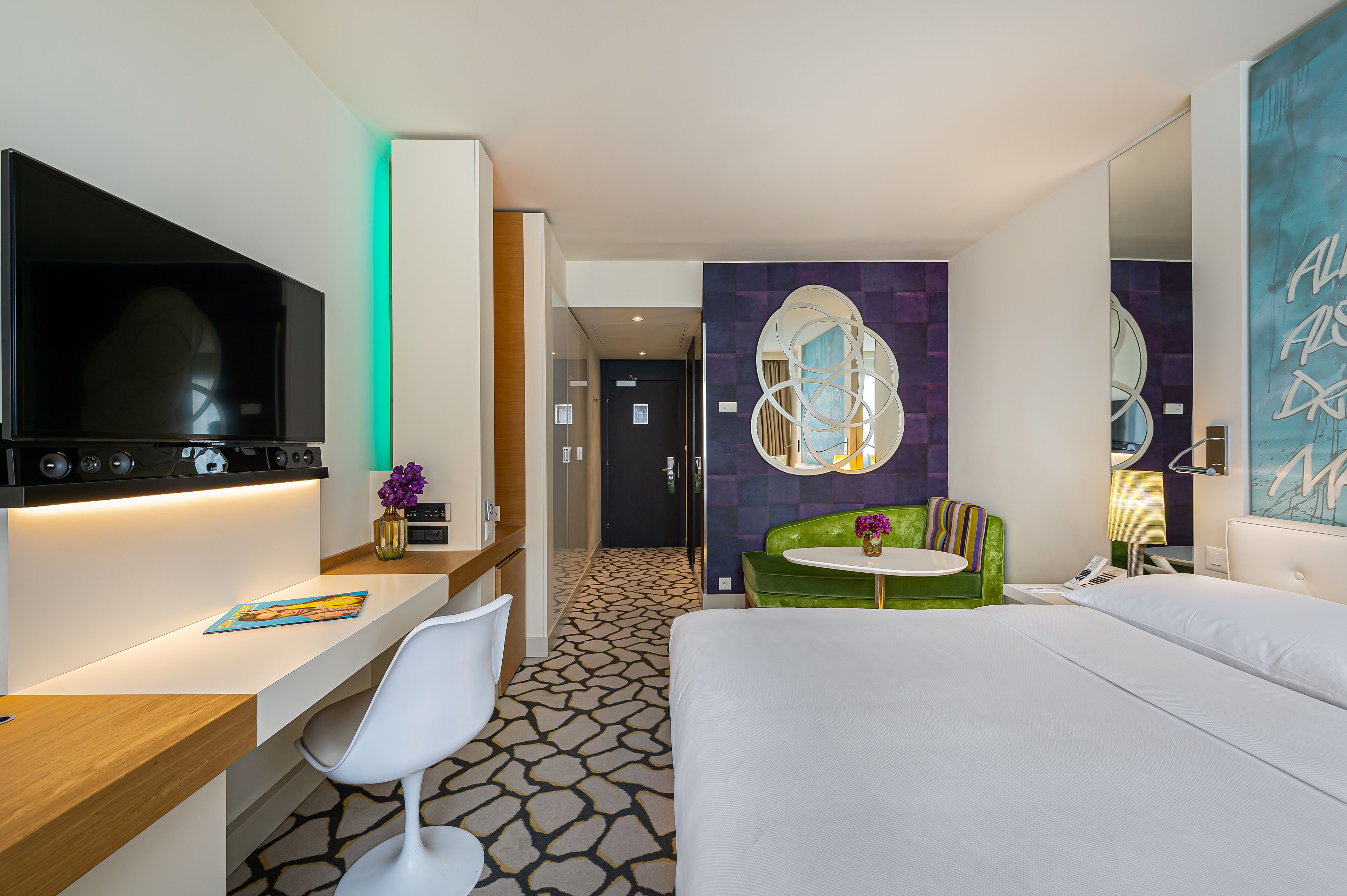 Une chambre d’hôtel moderne dotée d’un grand lit, d’un bureau avec un téléviseur à écran plat, d’un fauteuil de couleur vive et d’une petite table ronde.