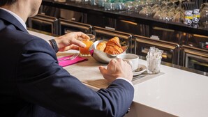 Un hombre sentado en la barra, con un croissant y un vaso de jugo de naranja. La barra tiene estantes con diferentes botellas y vasos.