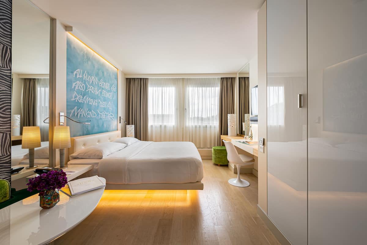 A modern hotel room with a large bed, bedside tables, a desk, and a large mirror.