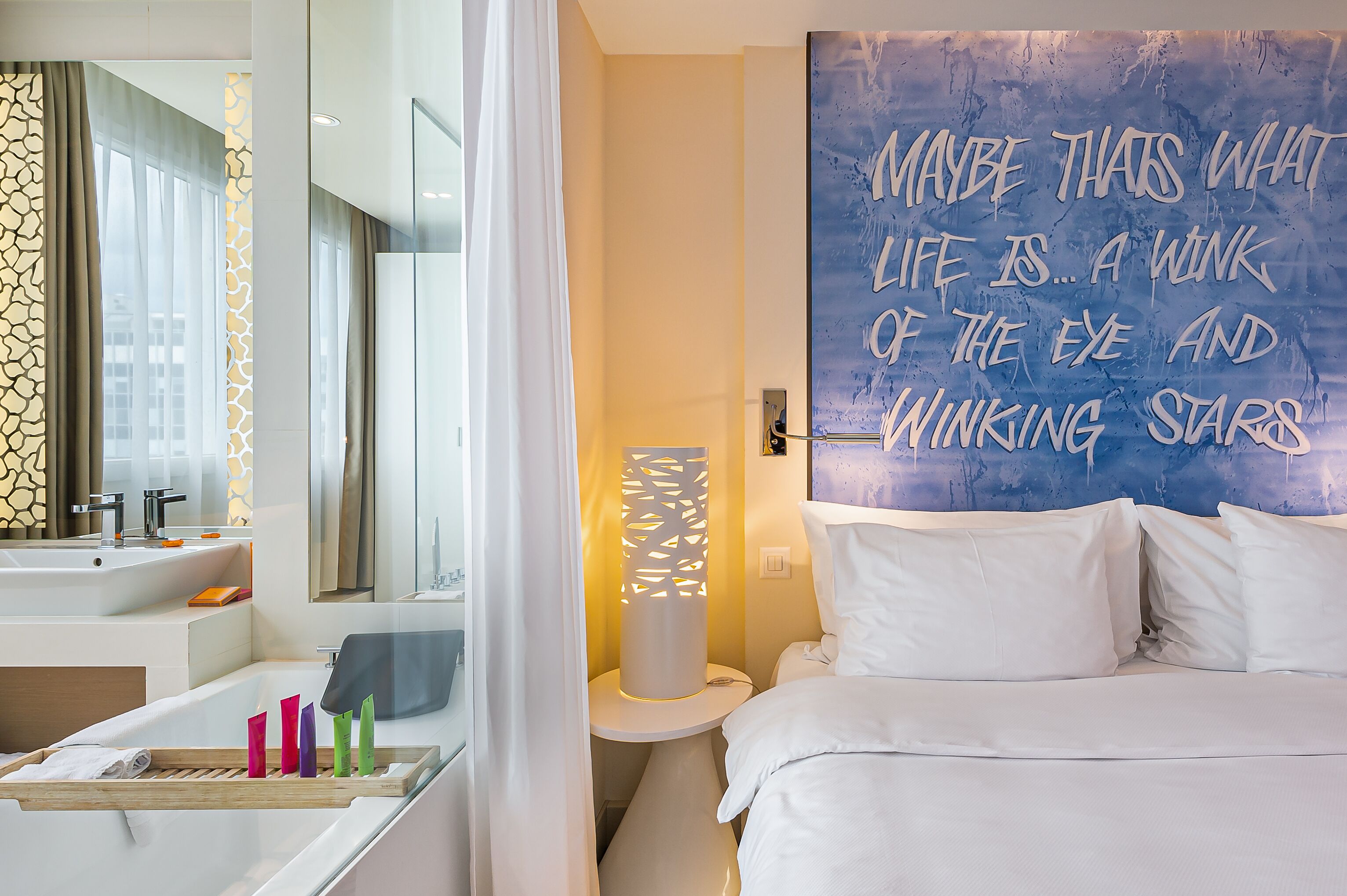 A modern hotel room with a large mirror, a bedside table with a lamp, and a wall with a motivational quote.