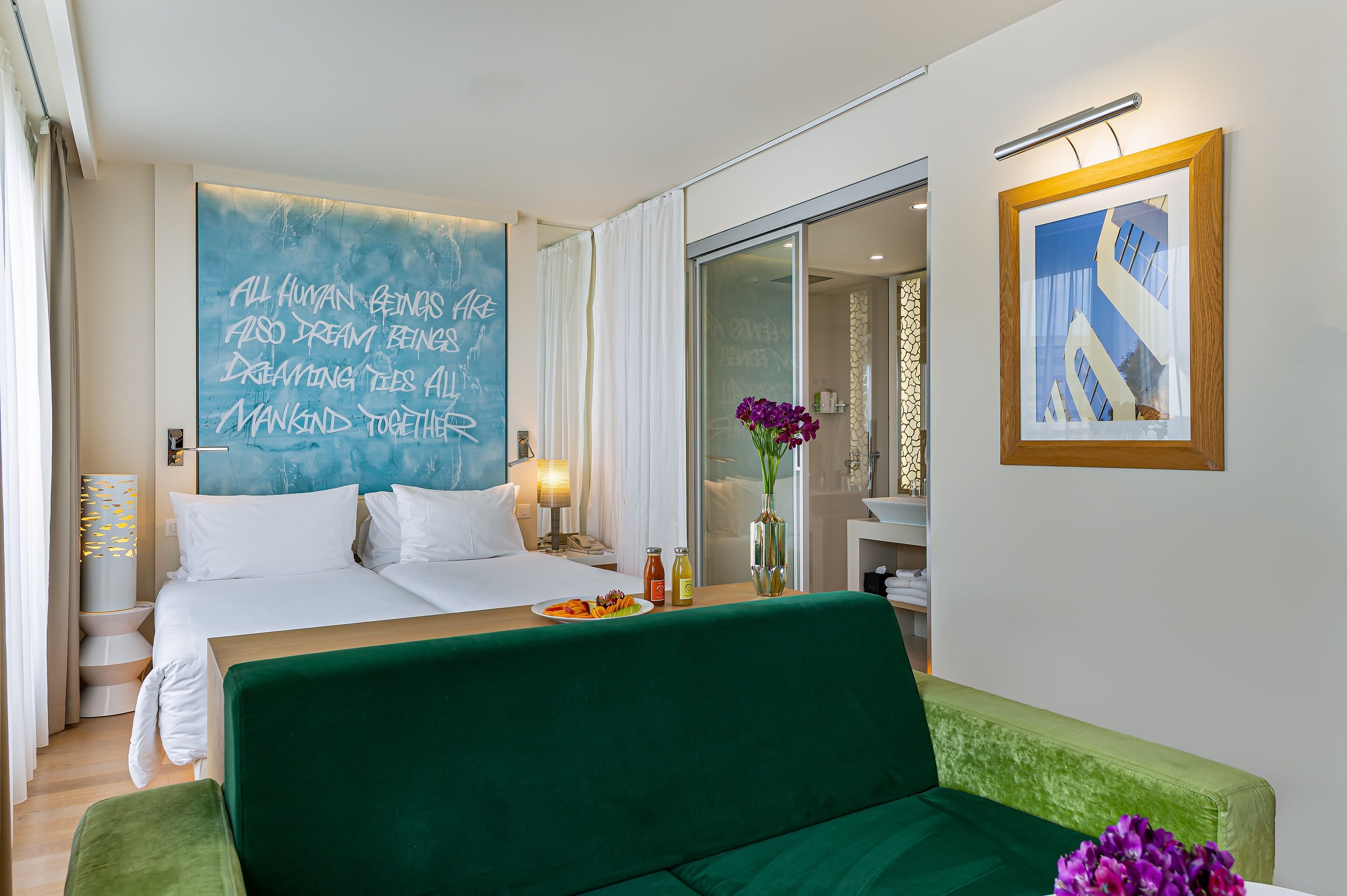 Habitación de hotel con una cama grande, un sofá verde, una mesa de comedor con desayuno, un baño con puerta de vidrio y una cita motivacional en la pared.