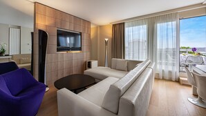 Habitación de hotel moderna con una televisión de pantalla plana grande montada en una pared de paneles de madera, un sofá color beige, un sillón morado, una mesita redonda y un balcón con vistas a la ciudad.
