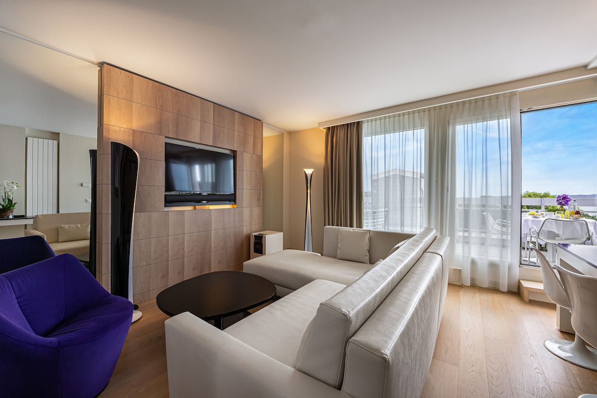 A modern hotel room with a large flat-screen TV mounted on a wooden panel wall, a beige sofa, a purple armchair, a small round table, and a balcony with a view of the city.